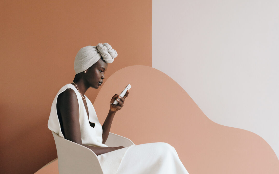 Side view of confident young black female model in trendy total white outfit and traditional turban sitting on chair and messaging on smartphone on two color background